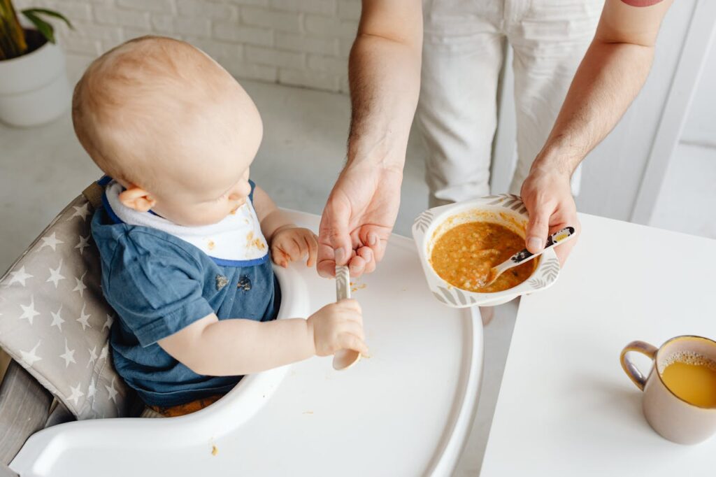 Rozszerzanie diety niemowlaka: mężczyzna próbuje nakarmić zupą dziecko siedzące na krzesełku do karmienia.