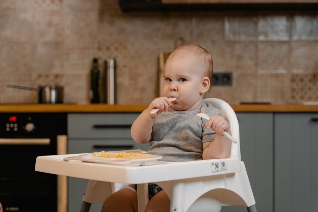 Dziecko siedzi w krzesełku do karmienia i spożywa posiłek. Trzyma łyżeczkę z jedzeniem w buzi. W tle nowoczesna kuchnia.