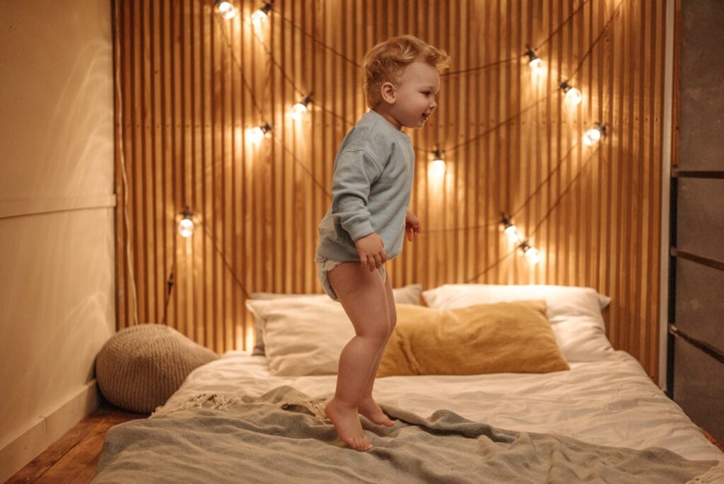 dziecko skaczące po łóżku. Dziecko ma pieluszkę i bluzkę z długim rękawem. W tle drewniana ściana z lampkami dekoracyjnymi.