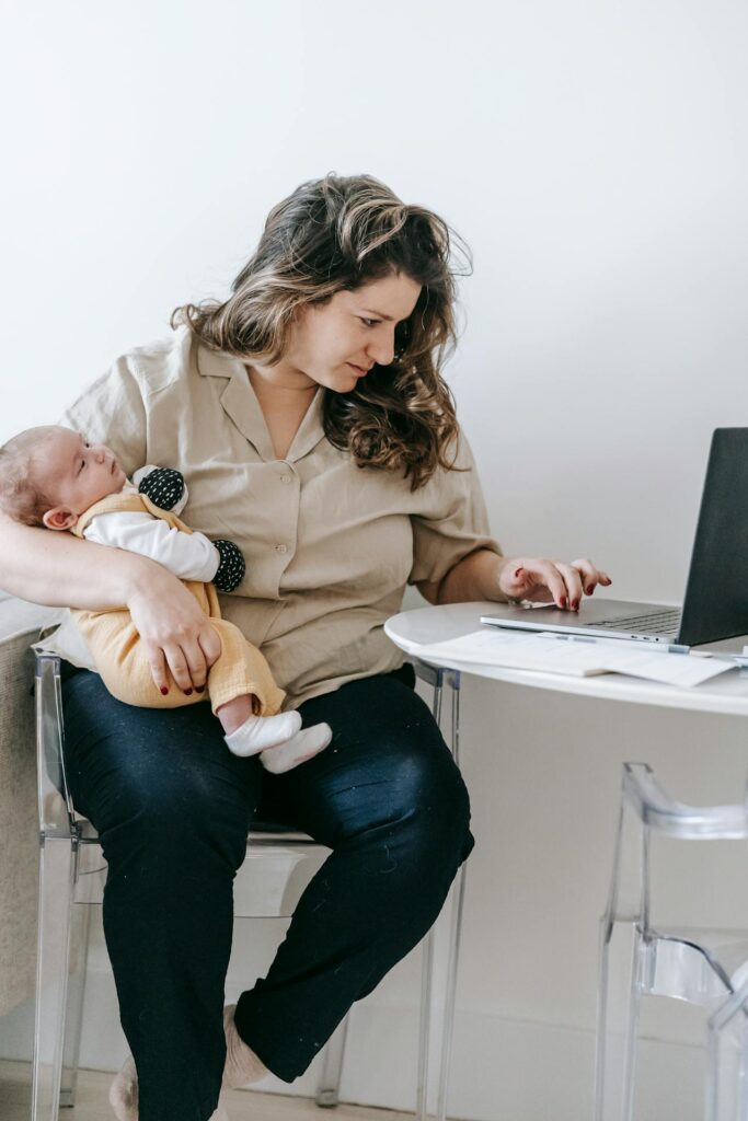 powrót do pracy po urlopie macierzyńskim: mama siedzi na krześle z dzieckiem na rękach i pracuje na laptopie