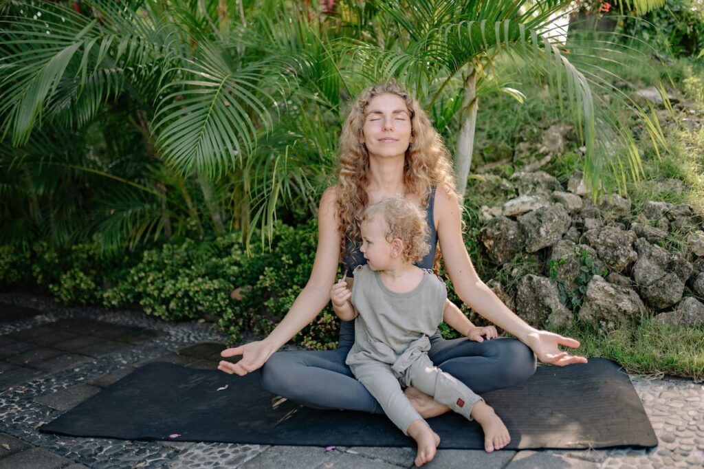 Kobieta z dzieckiem na kolanach ćwiczy jogę. W tle palmy i inne rośliny.