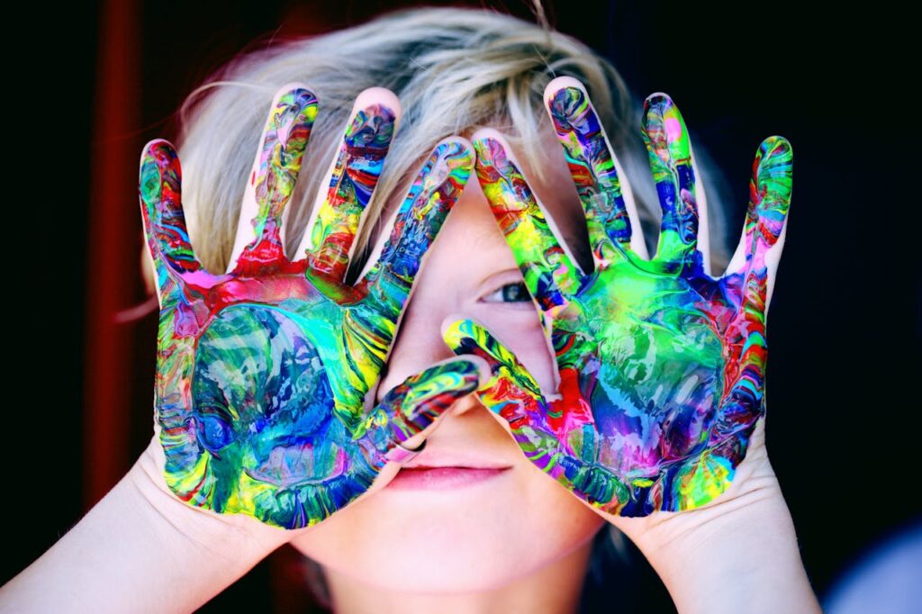 dziecko pozuje z wyciągniętymi przed twarzą dłońmi, które są pomalowane farbkami na różne kolory. Wzory na dłoniach ukłądają się w obrazki przedstawiające dzikie zwierzęta