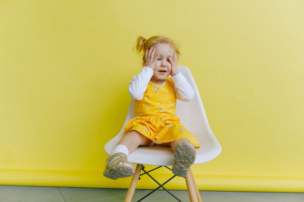 dziewczynka siedzi w żółtej sukience na białym krześle i trzyma się za głowę