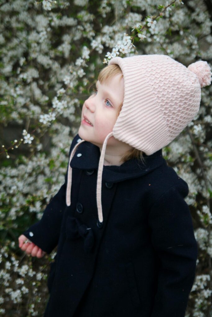 poradnik, jak ubierać dziecko wiosną. Dziecko w czarnym płaszczyku i jasnoróżowej czapce na tle drzewa z kwiatami