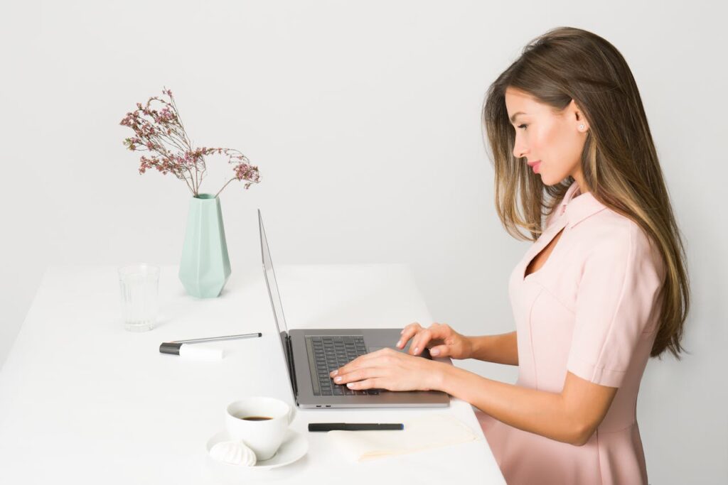 elegancka kobieta pracuje zdalnie przy laptopie, który znajduje się na białym biurku. Na biurku stoją nowoczesny wazon z kwiatem i szklanka z wodą.