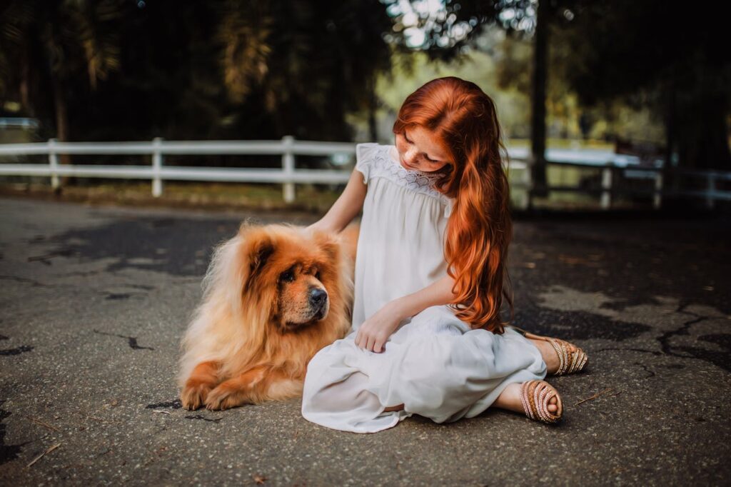 rudowłosa dziewczynka siedzi na ziemi i głaszcze psa rasy chow chow