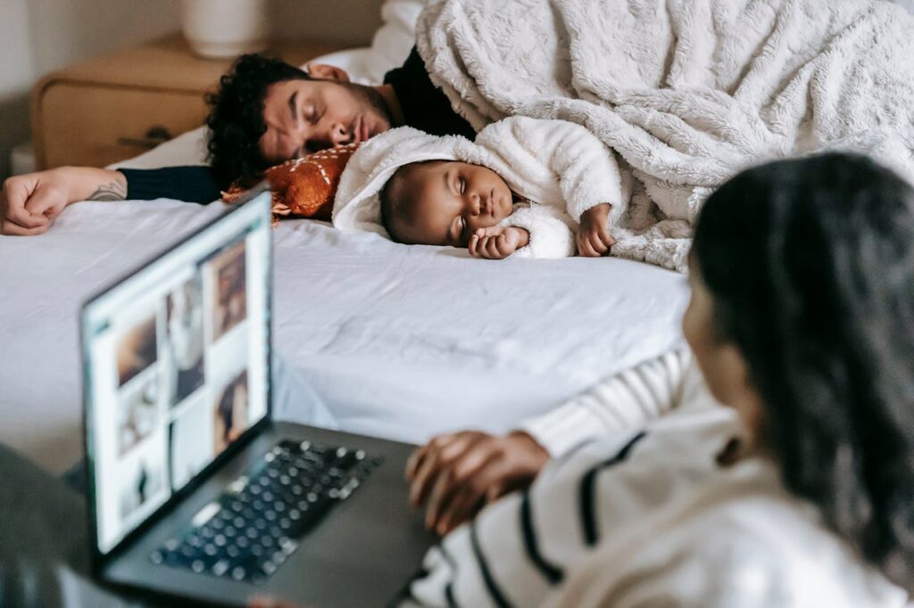 Mama pracuje przy laptopie w sypialni, a dziecko z tatą śpią na łóżku.